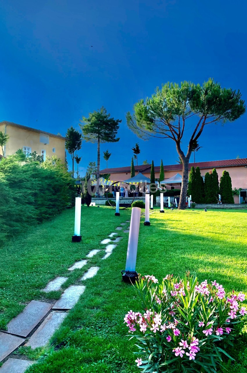 Pelouse avec chemin en dalles, colonnes lumineuses colorées, arbres et bâtiment en arrière-plan sous ciel bleu.