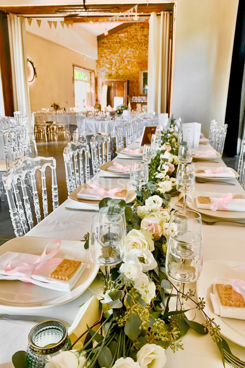 Table décorée pour un événement avec nappes blanches, fleurs, rubans roses et chaises transparentes dans une salle lumineuse.