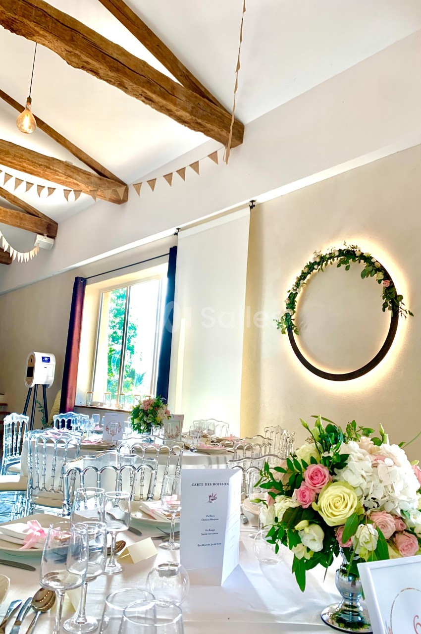 Salle de réception lumineuse décorée pour un mariage, avec tables dressées, guirlandes et couronne florale murale.