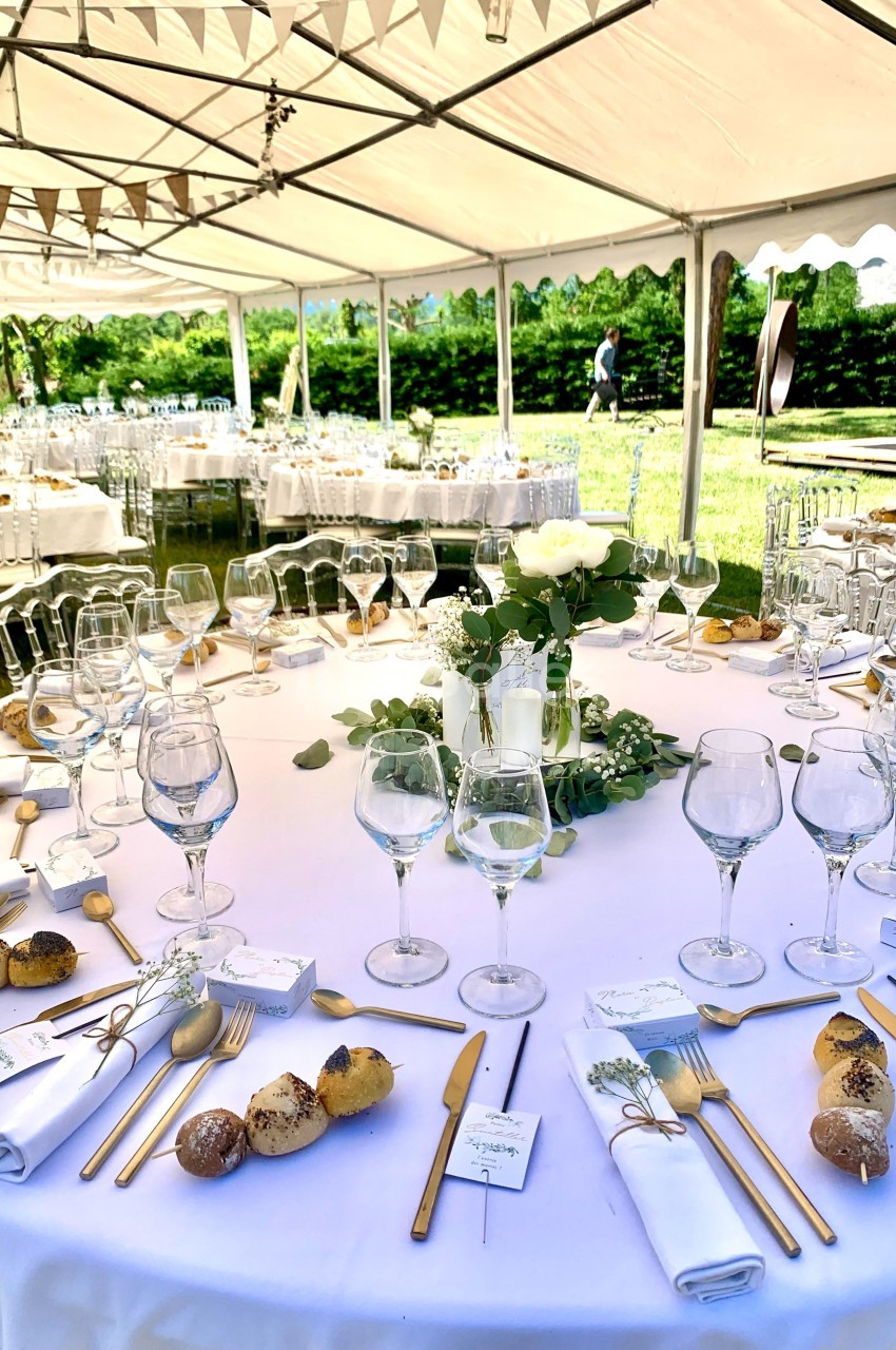 Table dressée pour un repas en extérieur sous une tente, avec vaisselle élégante, décorations florales et couverts dorés.
