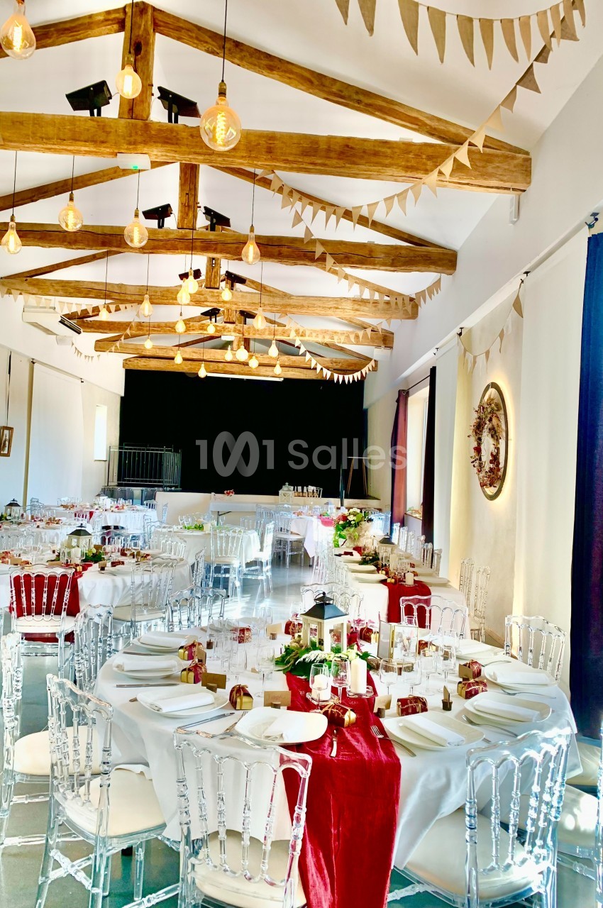 Salle de réception lumineuse avec tables rondes décorées de nappes blanches et rouges, guirlandes suspendues au plafond.