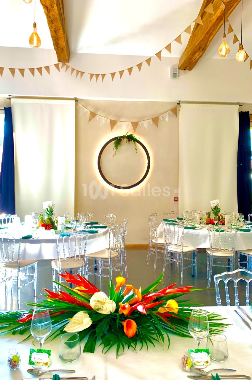 Salle de réception décorée avec des tables dressées, des fleurs tropicales et des guirlandes suspendues.