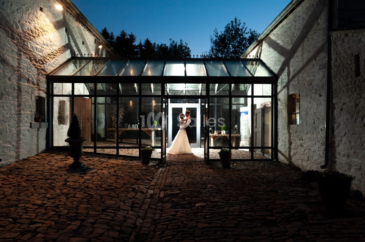Un couple en tenue de mariage s'embrasse sous une verrière illuminée, entouré de murs en pierre au crépuscule.