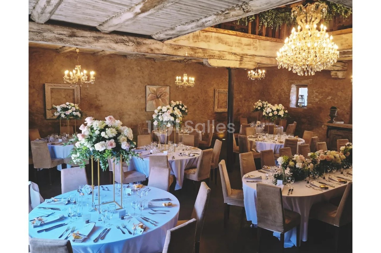 Salle de réception rustique avec tables rondes décorées de fleurs blanches et chandeliers suspendus.