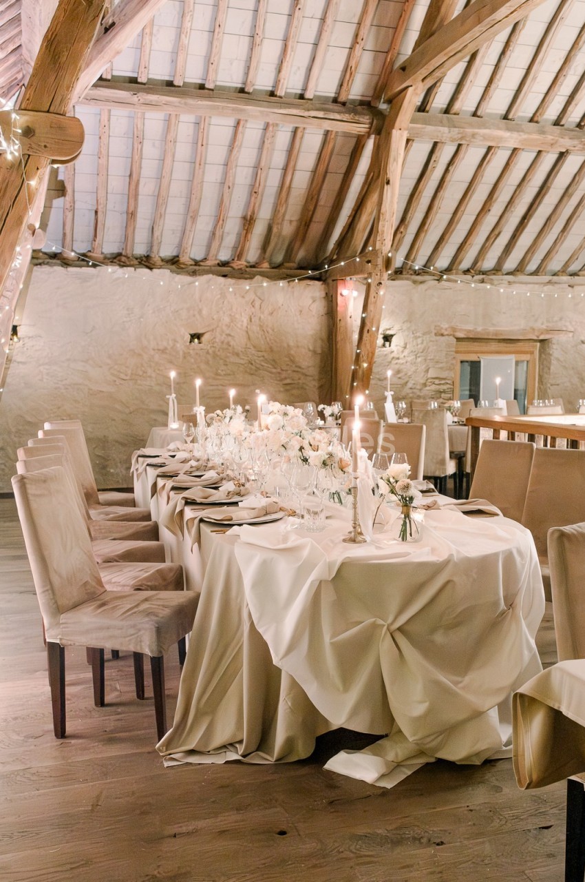 Table élégamment dressée avec nappes blanches, bougies et décorations florales, dans une salle en bois au plafond voûté.
