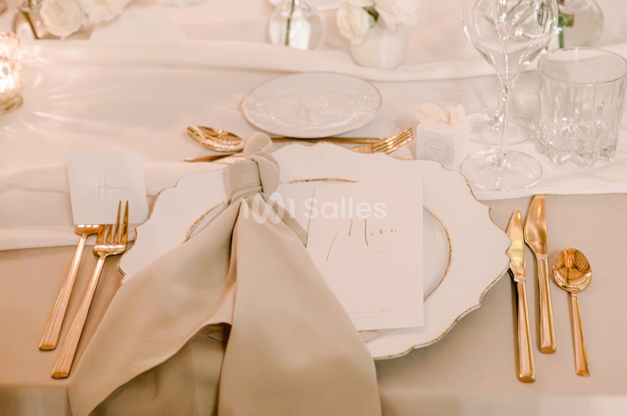 Table dressée avec une assiette blanche bordée d'or, couverts dorés, serviette nouée et menu posé dessus.