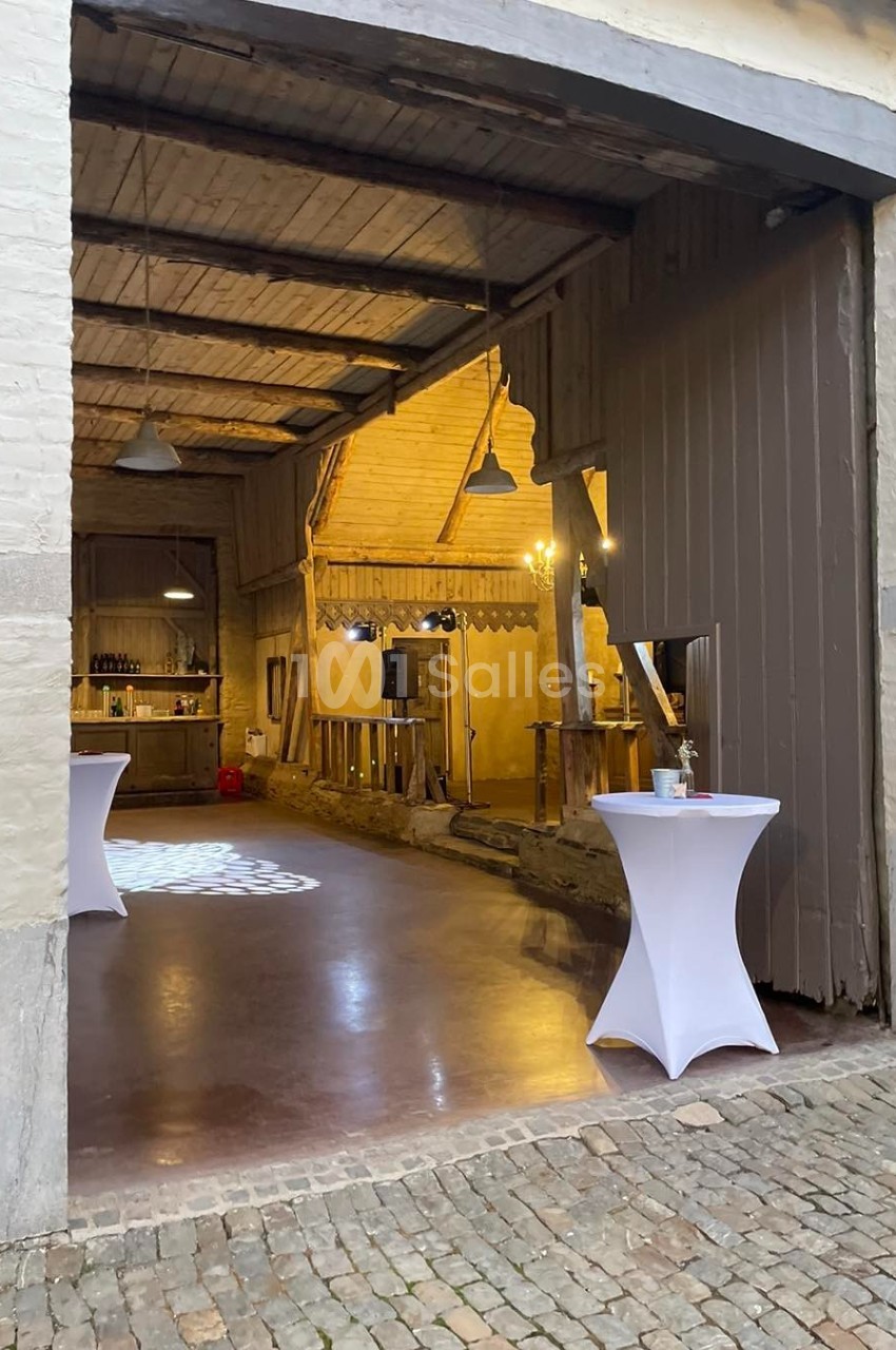 Entrée d'une grange aménagée pour un événement, avec tables hautes et éclairage intérieur chaleureux.