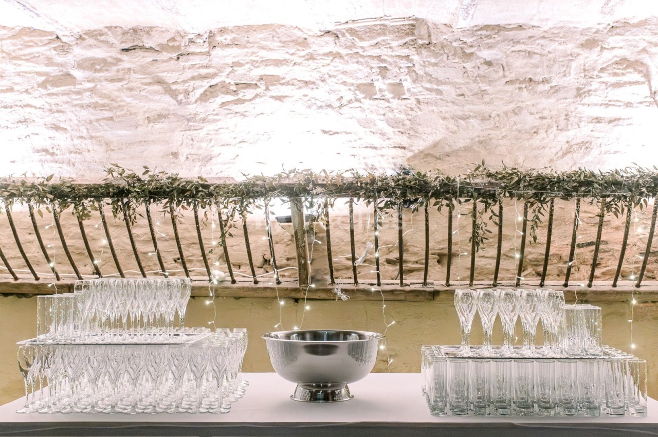 Table décorée avec des verres en cristal, une vasque en métal et une guirlande lumineuse sous un mur en pierre.