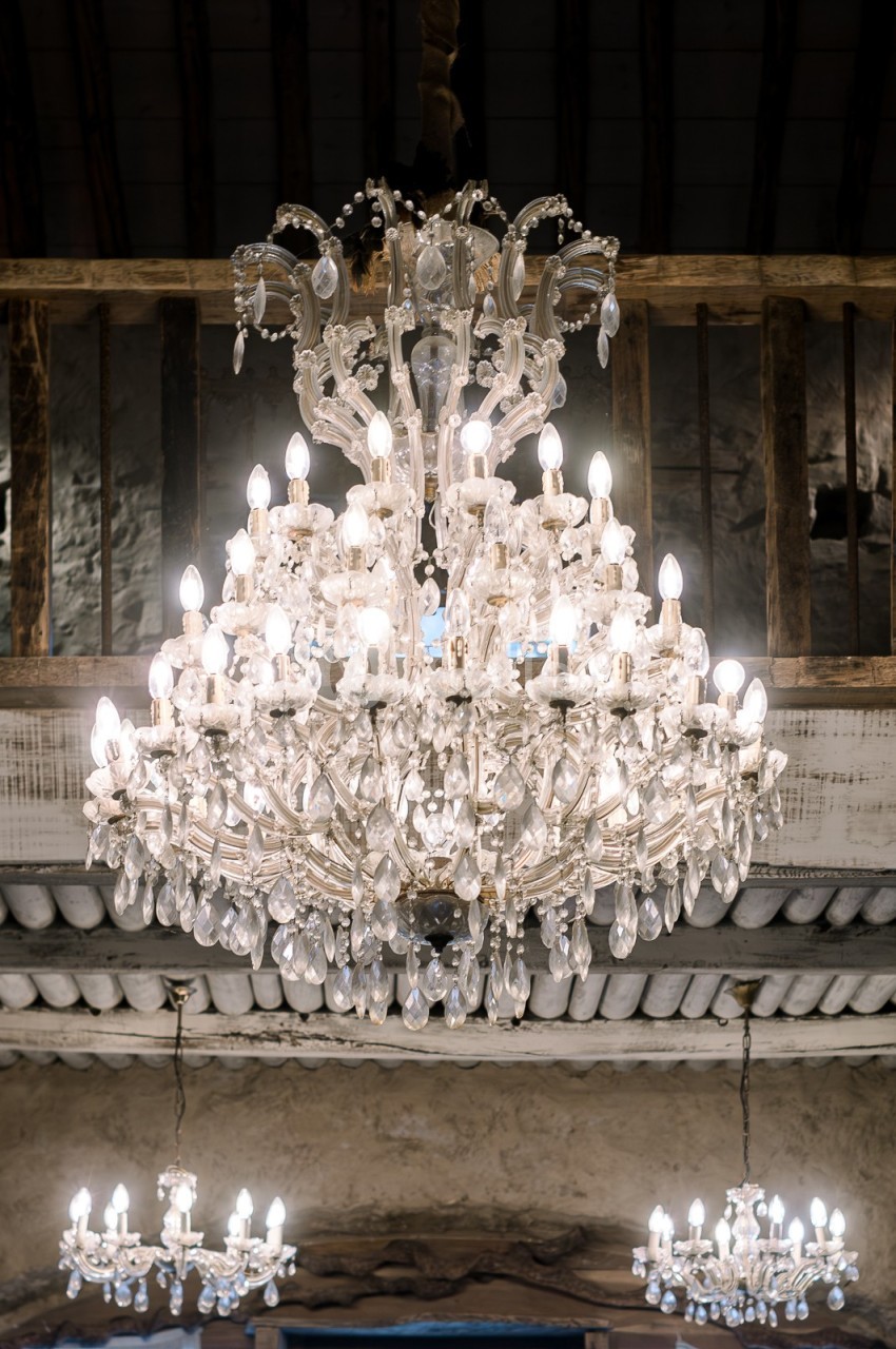 Lustre en cristal éclairé suspendu dans une pièce au plafond en bois et poutres apparentes.