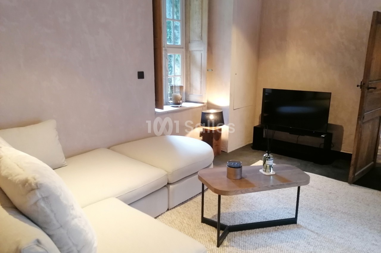 Salon avec canapé d'angle beige, table basse en bois, lampe, télévision et fenêtre laissant entrer la lumière naturelle.