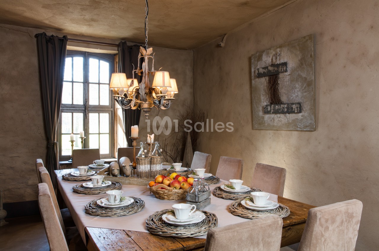Salle à manger rustique avec table en bois dressée, vaisselle blanche, panier de fruits et lustre suspendu.