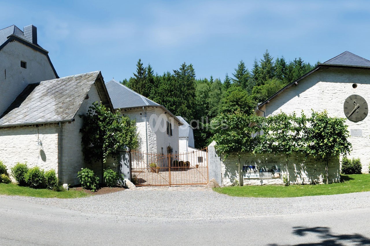 Portail en fer forgé entouré de bâtiments en pierre blanche avec toits en ardoise, bordés de végétation et d'arbres.