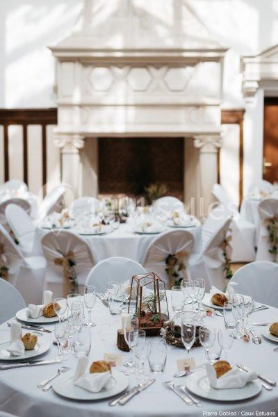 Tables rondes élégamment dressées avec nappes blanches, décorations florales et vaisselle, dans une salle lumineuse avec…