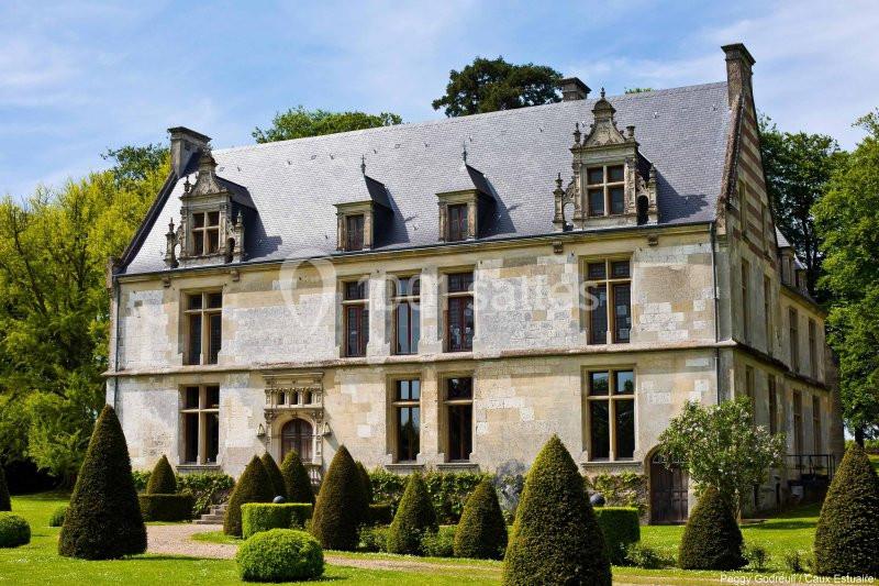 Façade d'un manoir en pierre avec toit en ardoise, entouré de buissons taillés et d'arbres dans un jardin.
