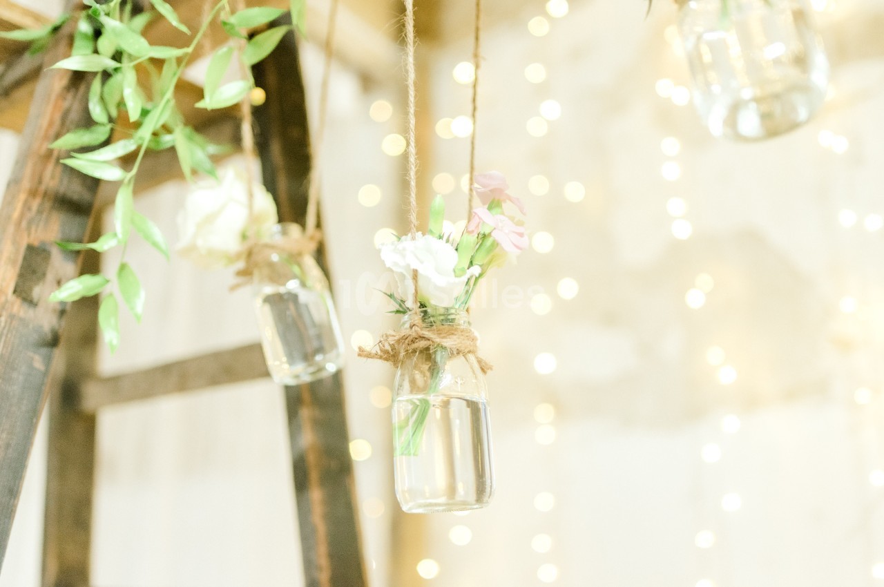 Petits vases suspendus en verre contenant des fleurs, accrochés à des cordes dans un décor lumineux et flou.