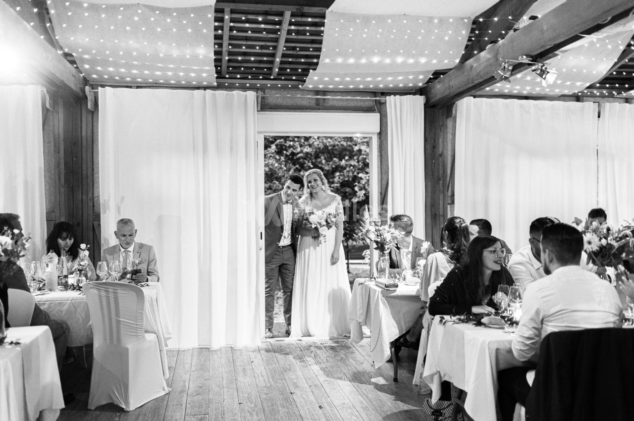 Un couple de mariés entre dans une salle de réception décorée, sous le regard des invités assis à des tables.