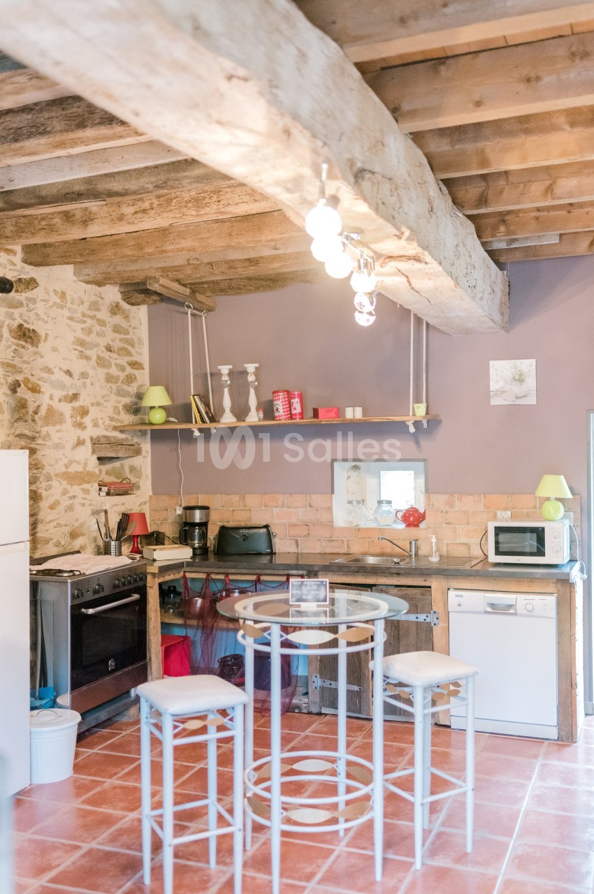 Cuisine rustique avec poutres apparentes, murs en pierre, électroménagers modernes et table ronde avec tabourets.