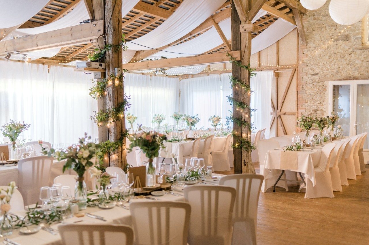 Salle de réception décorée avec des tables dressées, des fleurs, des guirlandes lumineuses et un plafond en tissu blanc.