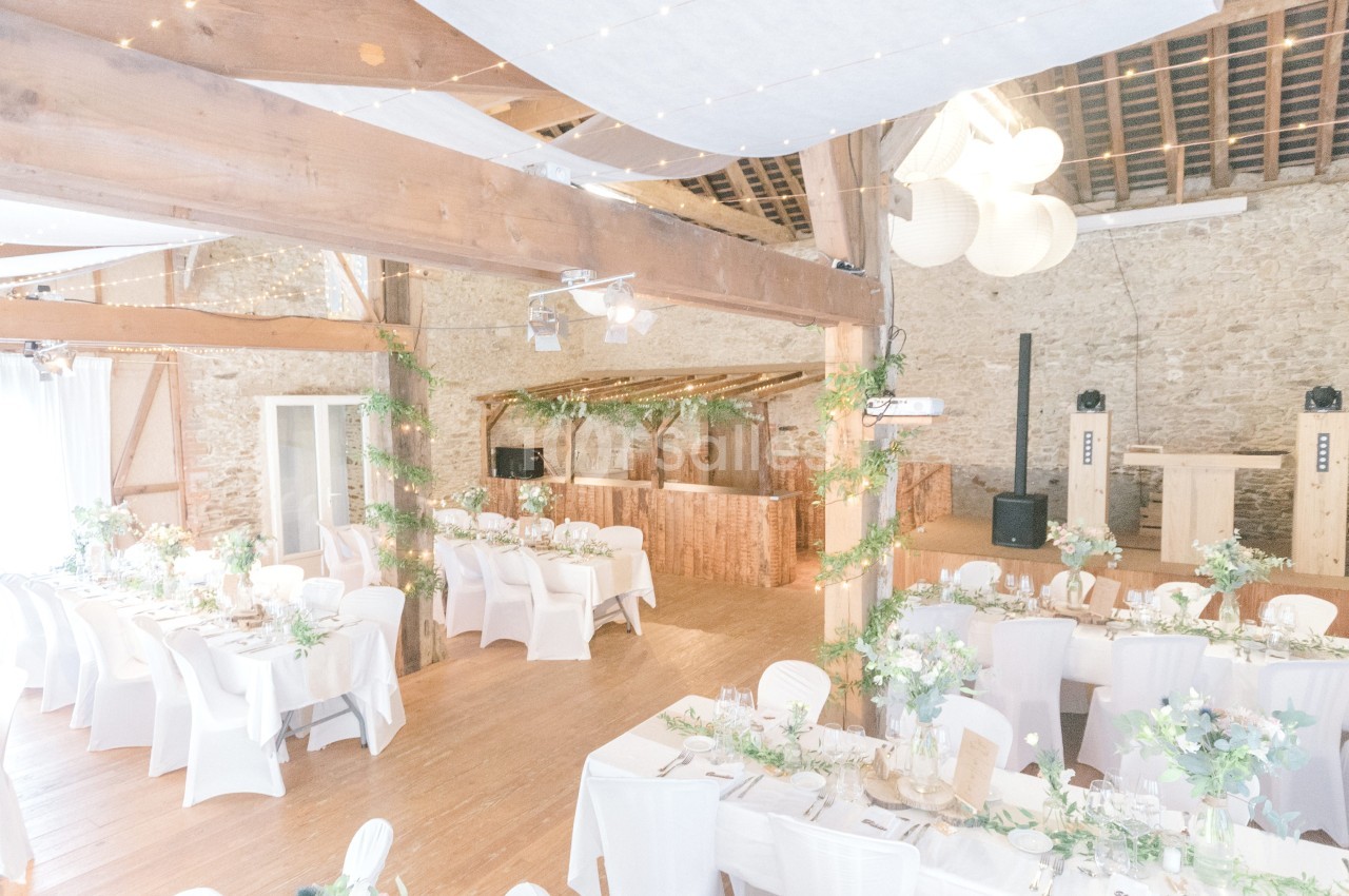 Salle de réception lumineuse avec tables décorées de fleurs, chaises blanches et guirlandes suspendues.