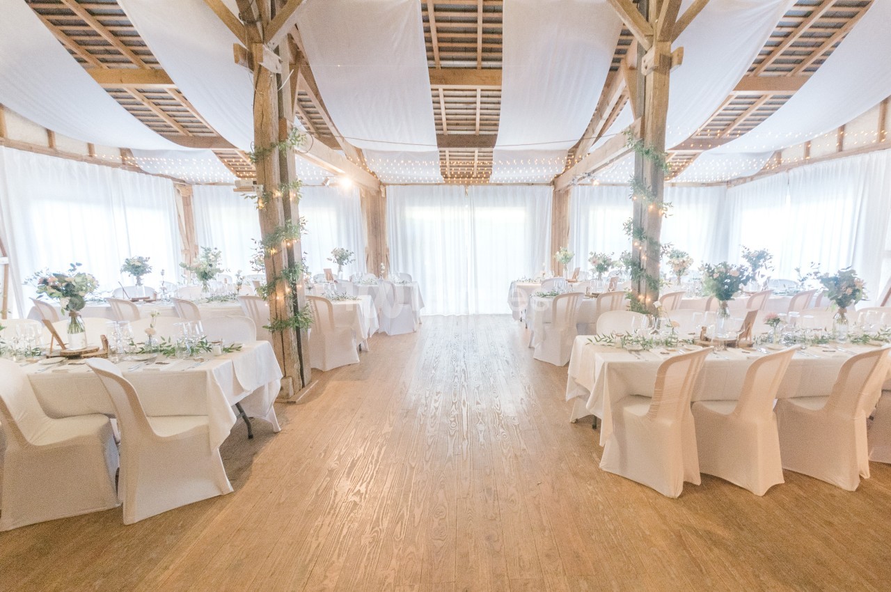 Salle de réception lumineuse décorée pour un événement, avec tables dressées et chaises blanches.