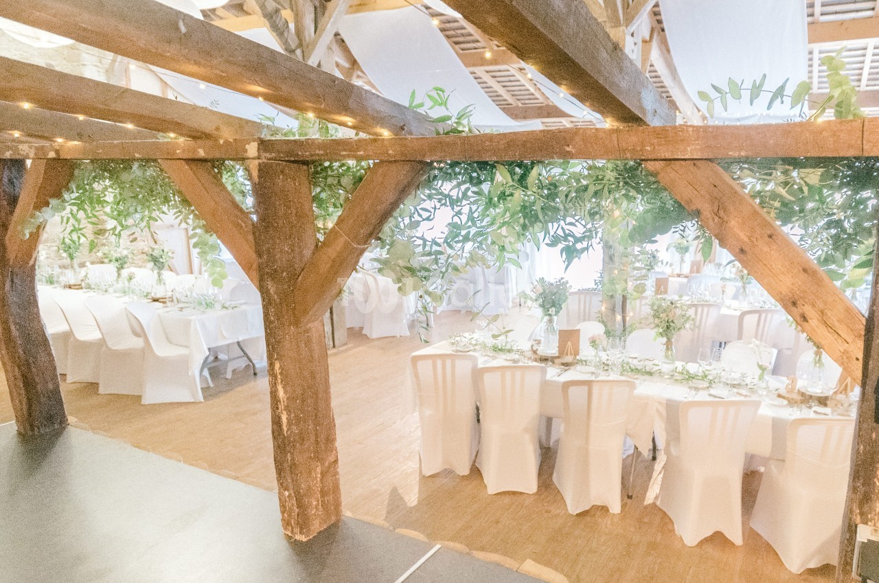 Salle de réception lumineuse avec tables décorées, chaises blanches et poutres en bois ornées de feuillage.