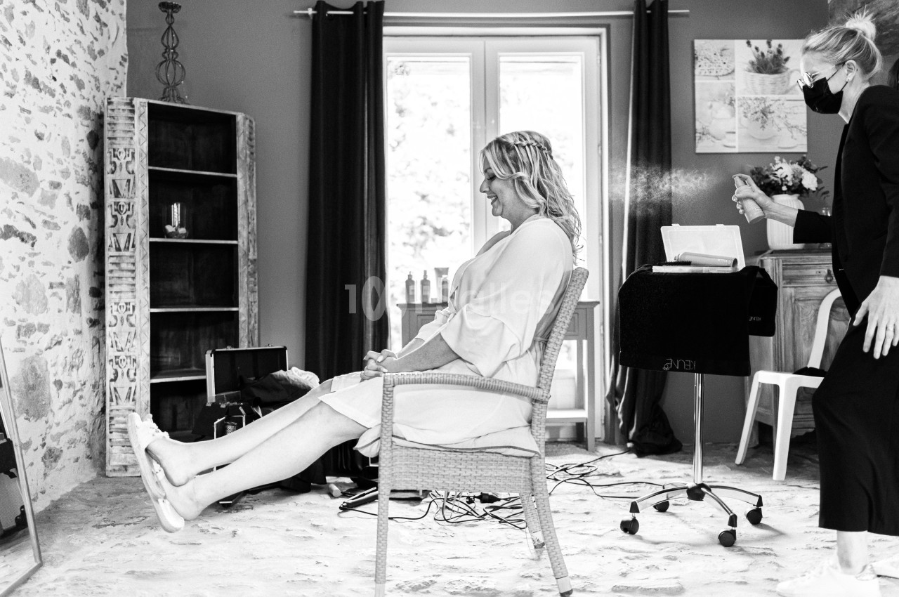 Une femme assise dans un fauteuil se fait coiffer dans une pièce lumineuse, avec une coiffeuse portant un masque.