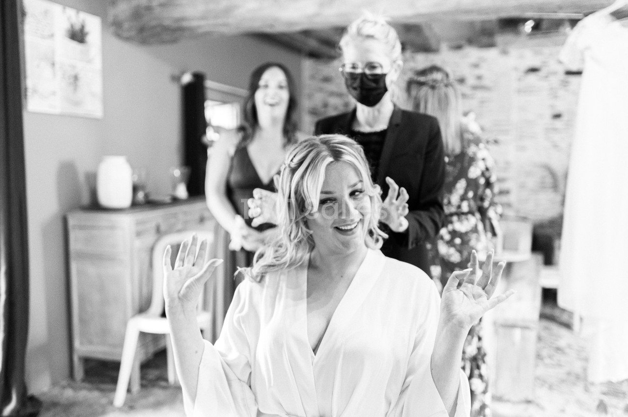 Une femme souriante en robe de préparation, entourée de personnes dans une pièce lumineuse au décor rustique.