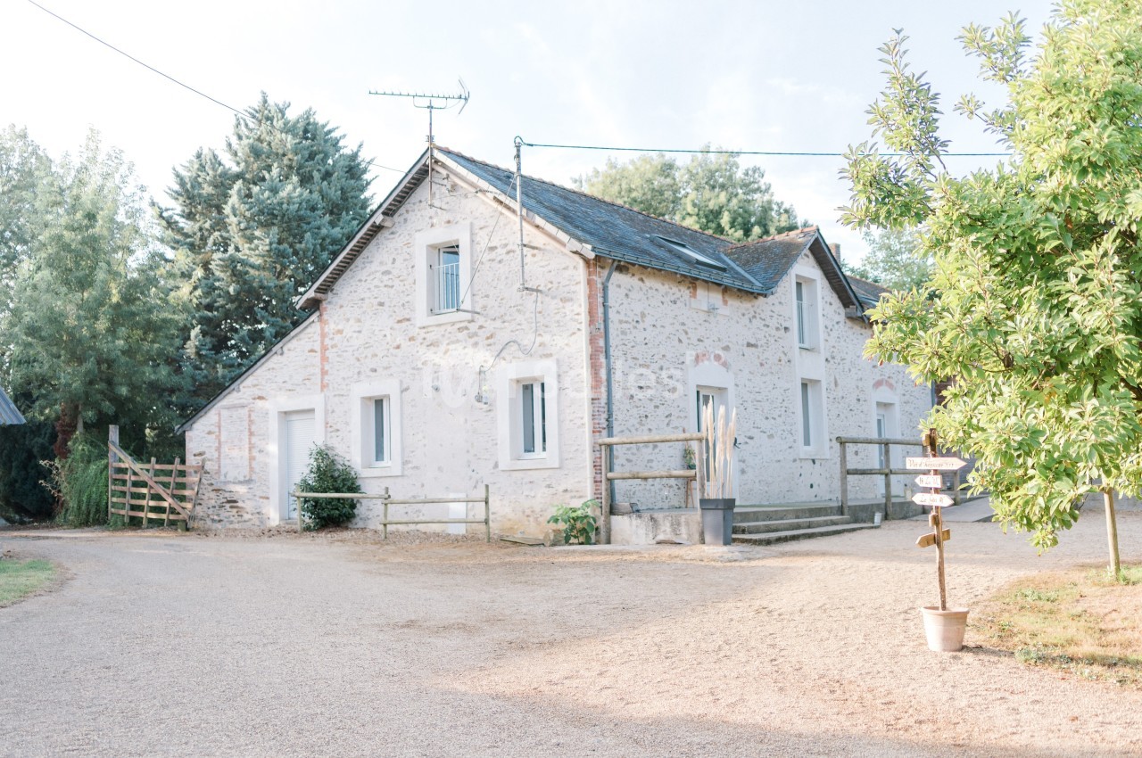 Maison en pierre blanche entourée de végétation, avec une cour en gravier et un panneau indicateur au premier plan.