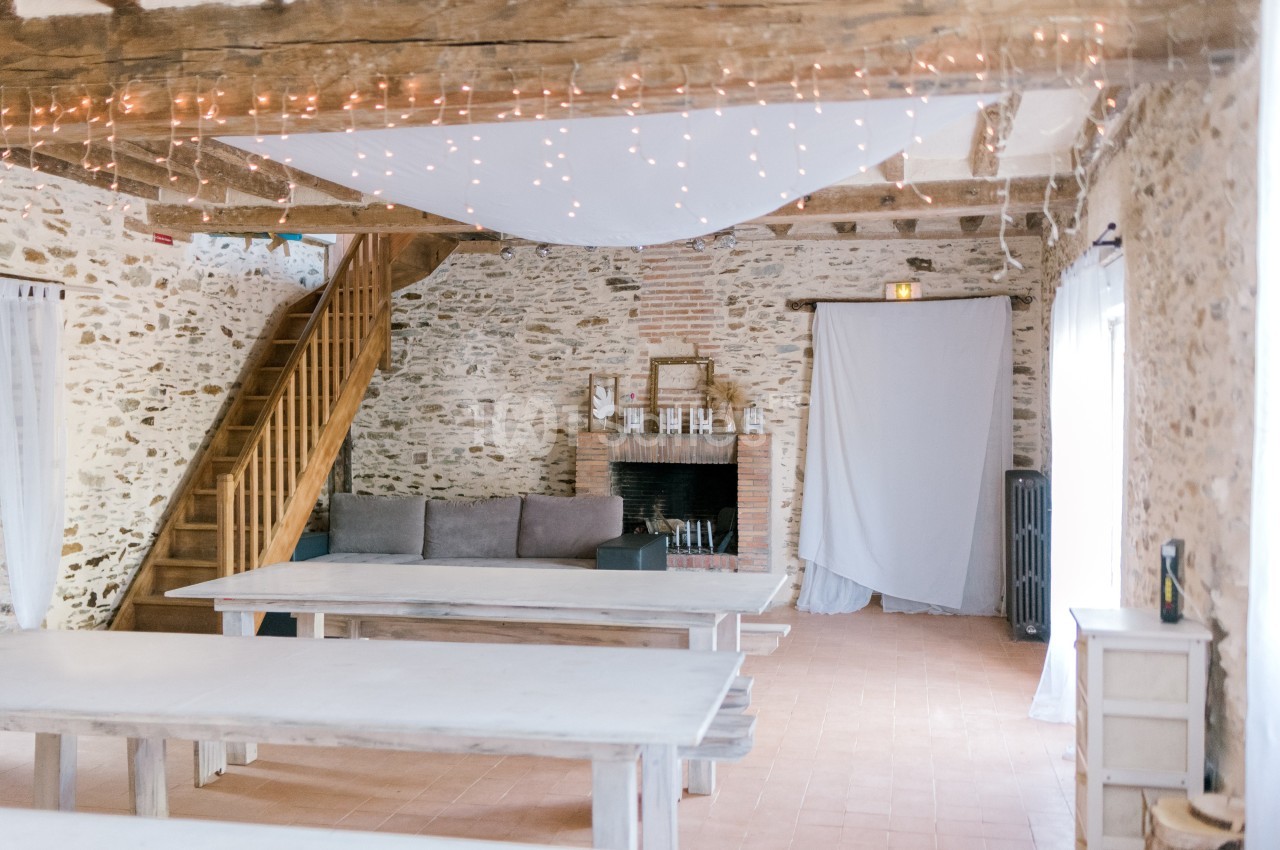 Salle lumineuse avec murs en pierre, poutres apparentes, tables en bois clair, canapé, cheminée et guirlandes lumineuses.
