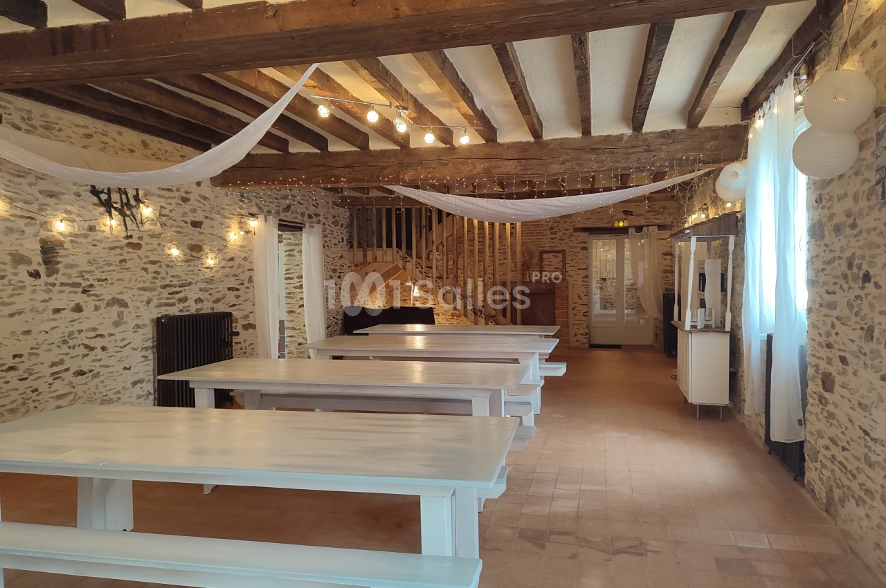 Salle rustique avec murs en pierre, poutres apparentes, grandes tables en bois peintes en blanc et guirlandes lumineuses.