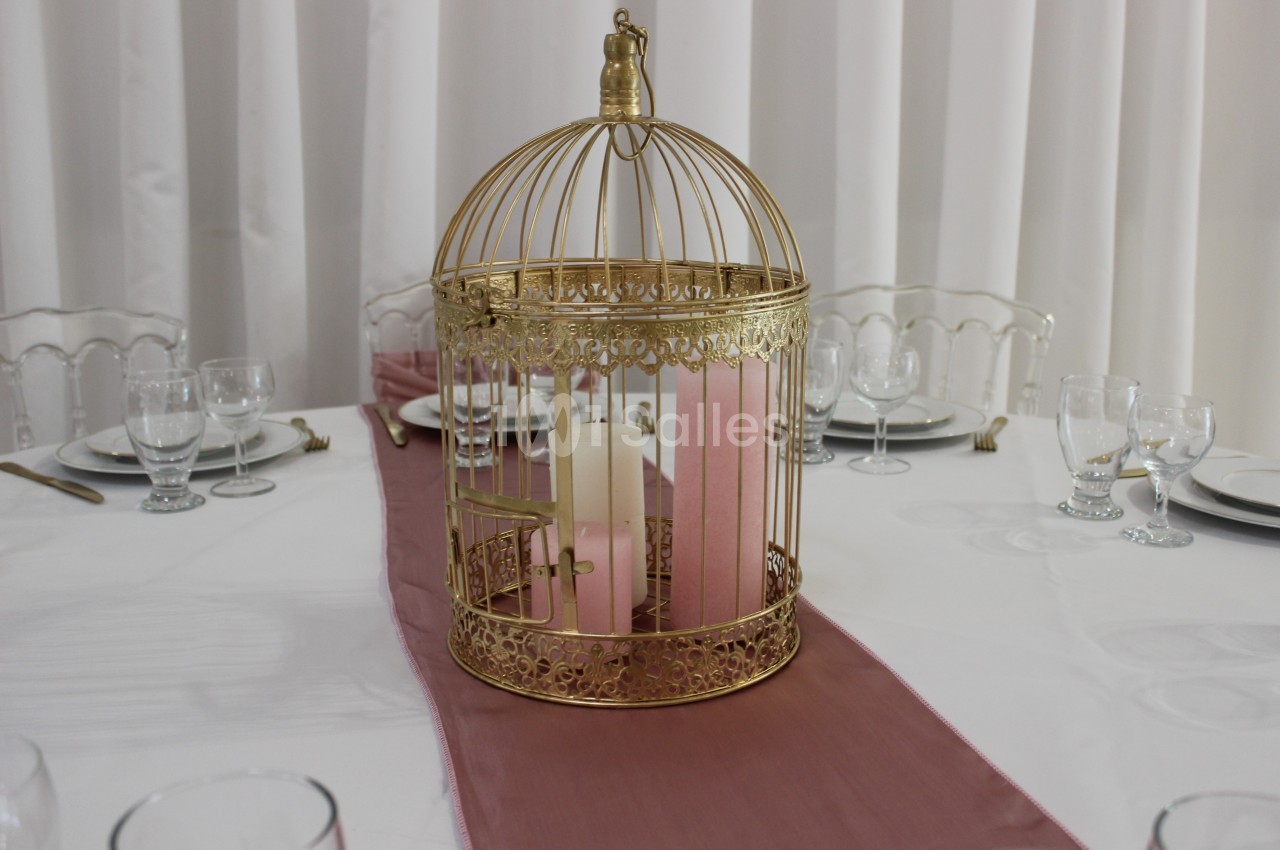 Centre de table avec une cage dorée contenant des bougies roses et blanches, posée sur un chemin de table rose.