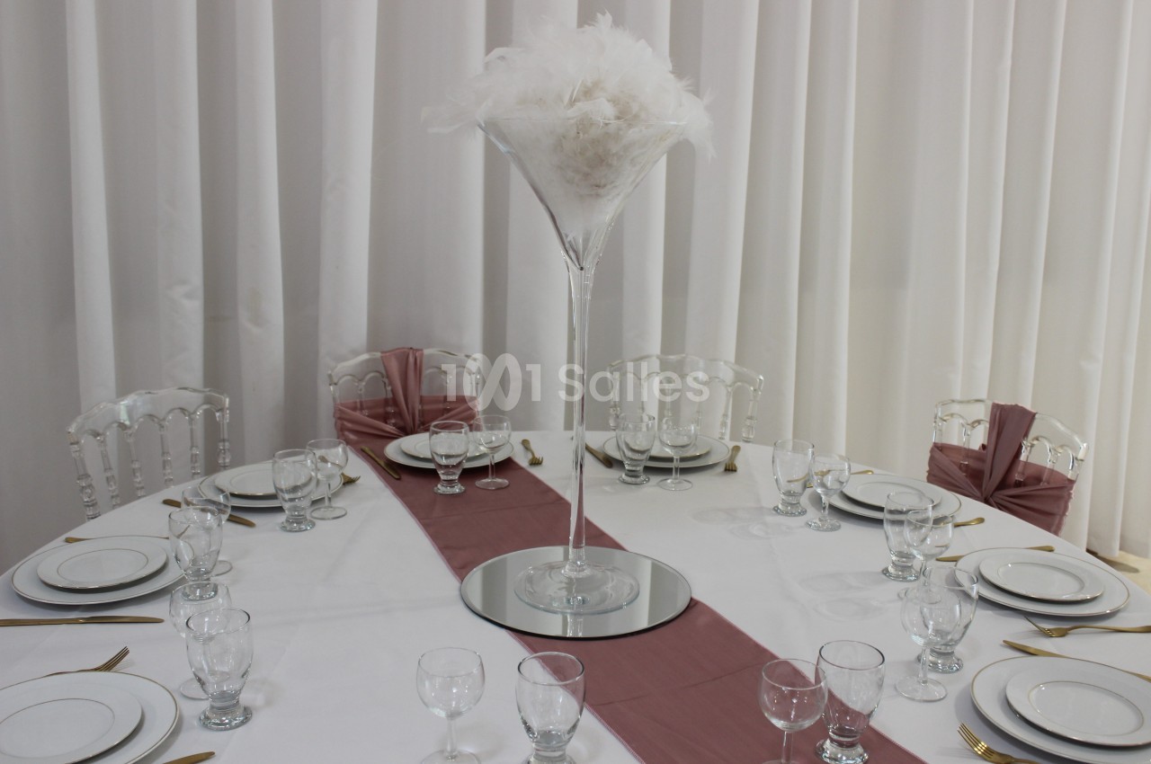 Table ronde dressée avec vaisselle blanche, couverts dorés, chemin de table rose et centre de table en plumes blanches.