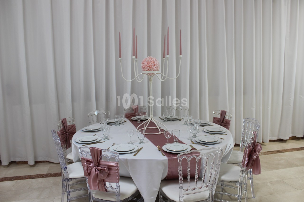 Table ronde élégamment dressée avec nappes blanches, chaises transparentes ornées de nœuds roses et chandelier central.