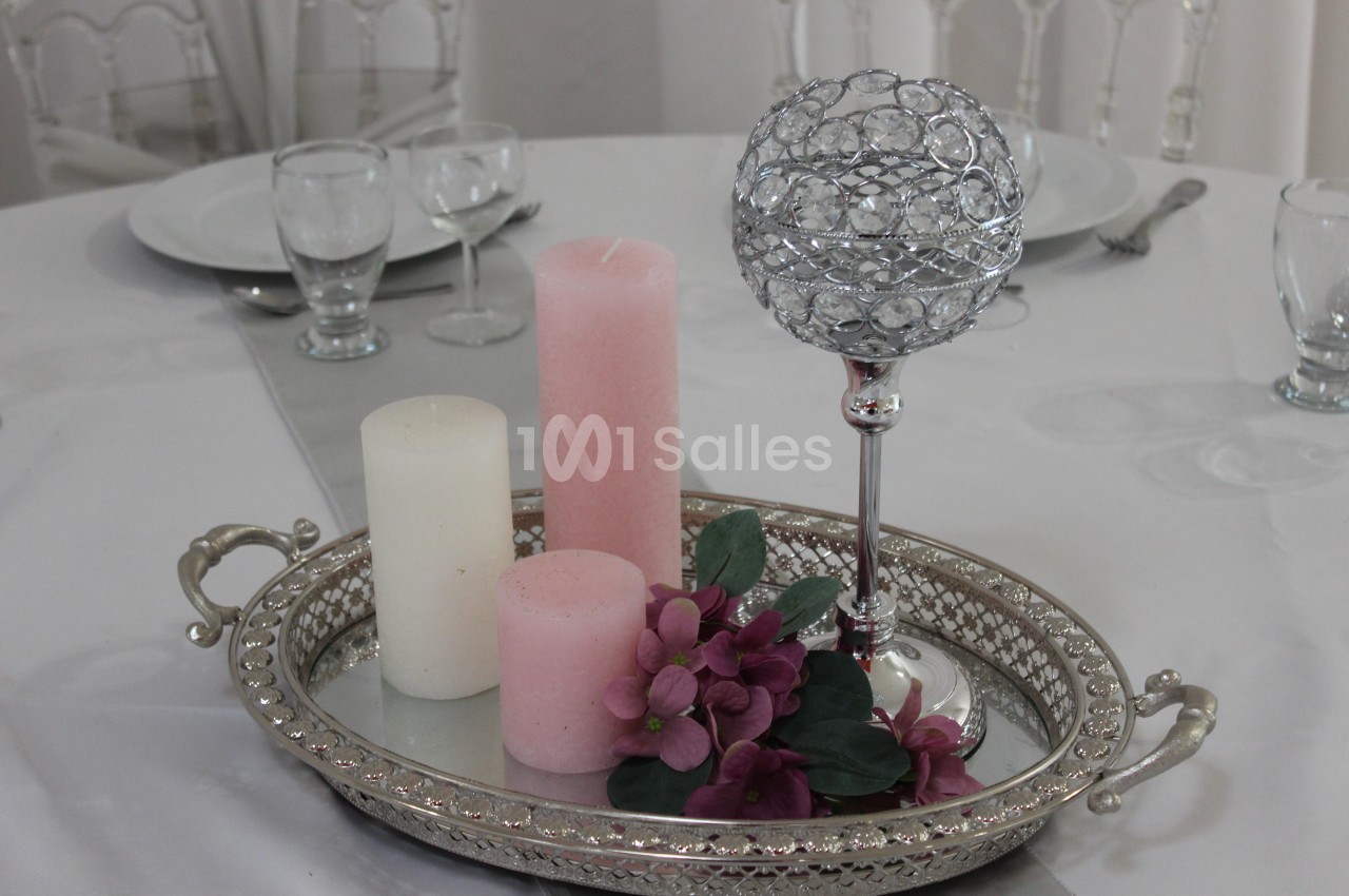 Centre de table avec bougies blanches et roses, fleurs violettes et support argenté décoratif sur une table dressée.
