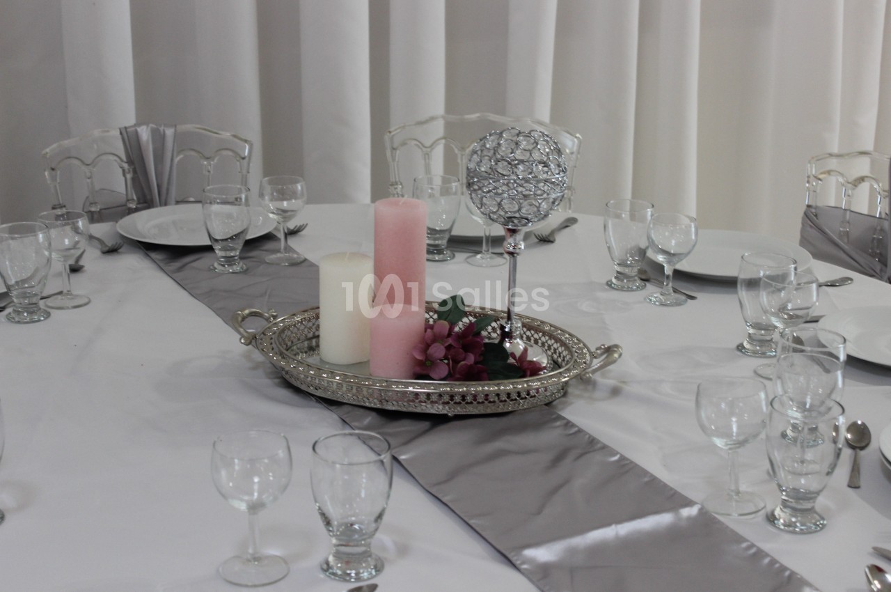 Table dressée avec nappe blanche, chemins de table gris, bougies et décoration florale sur un plateau argenté.