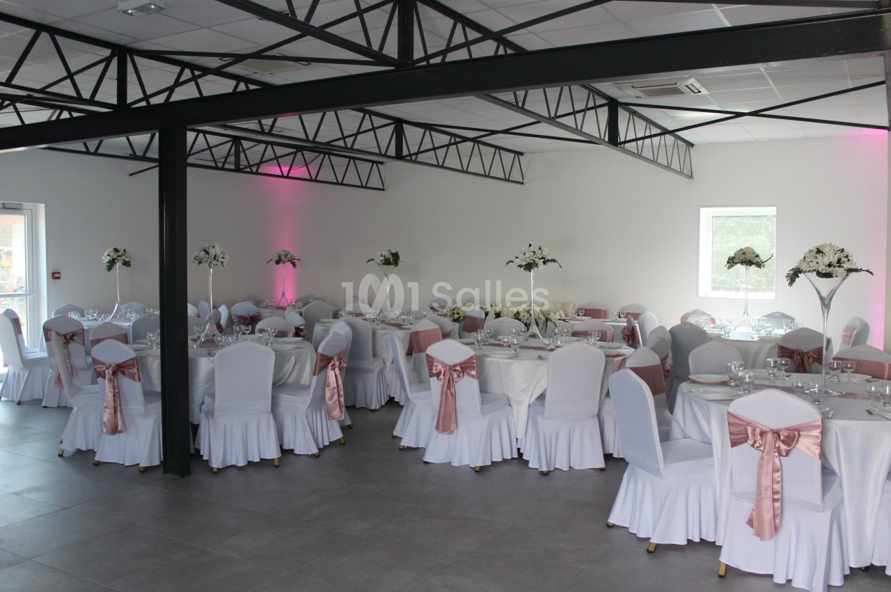Salle de réception décorée avec des tables rondes, nappes blanches, chaises ornées de nœuds roses et centres de table…