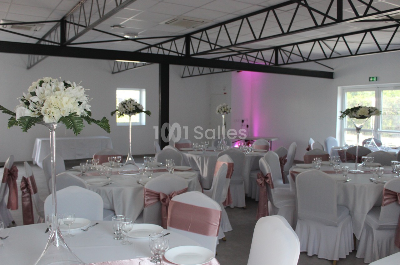 Salle de réception décorée avec des tables rondes, nappes blanches, chaises ornées de rubans roses et centres de table…