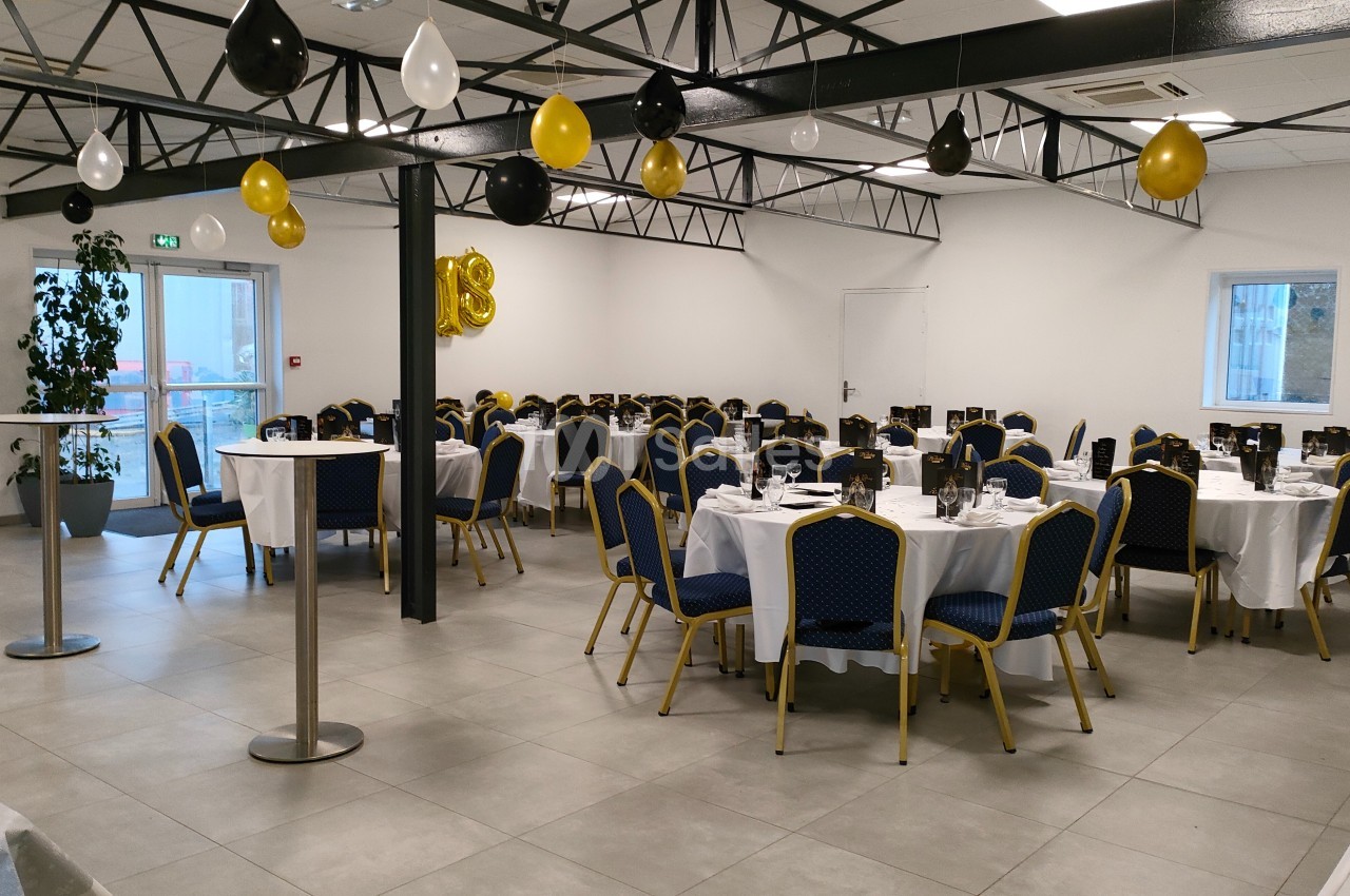 Salle de réception décorée avec des ballons noirs, blancs et dorés, tables rondes dressées avec nappes blanches et chaises…