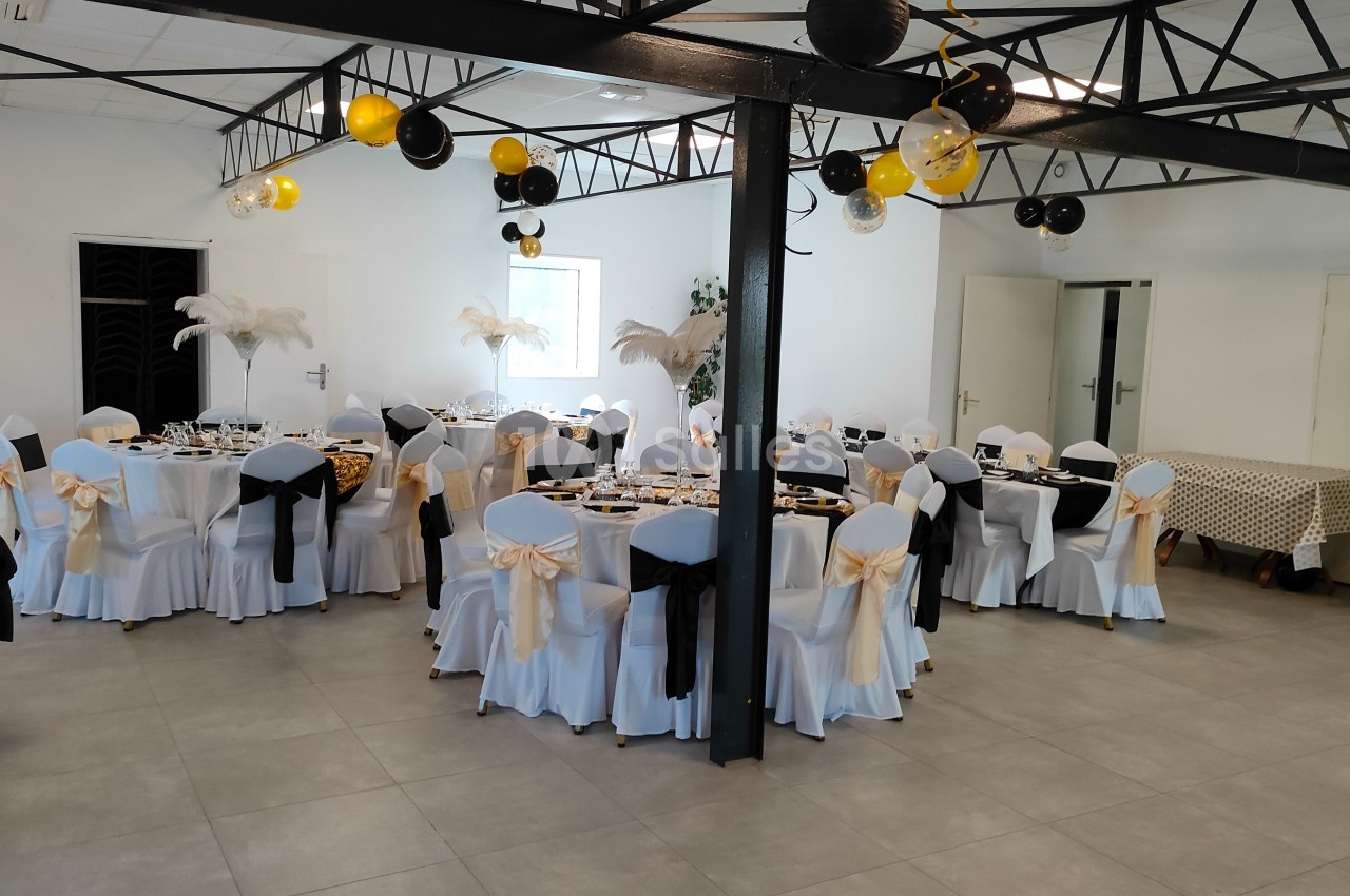 Salle de réception décorée avec des tables rondes, nappes blanches, chaises ornées de nœuds noirs et dorés.