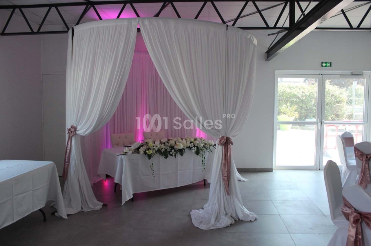 Décoration de salle avec arche en tissu blanc, table ornée de fleurs blanches et roses, éclairage rose en arrière-plan.