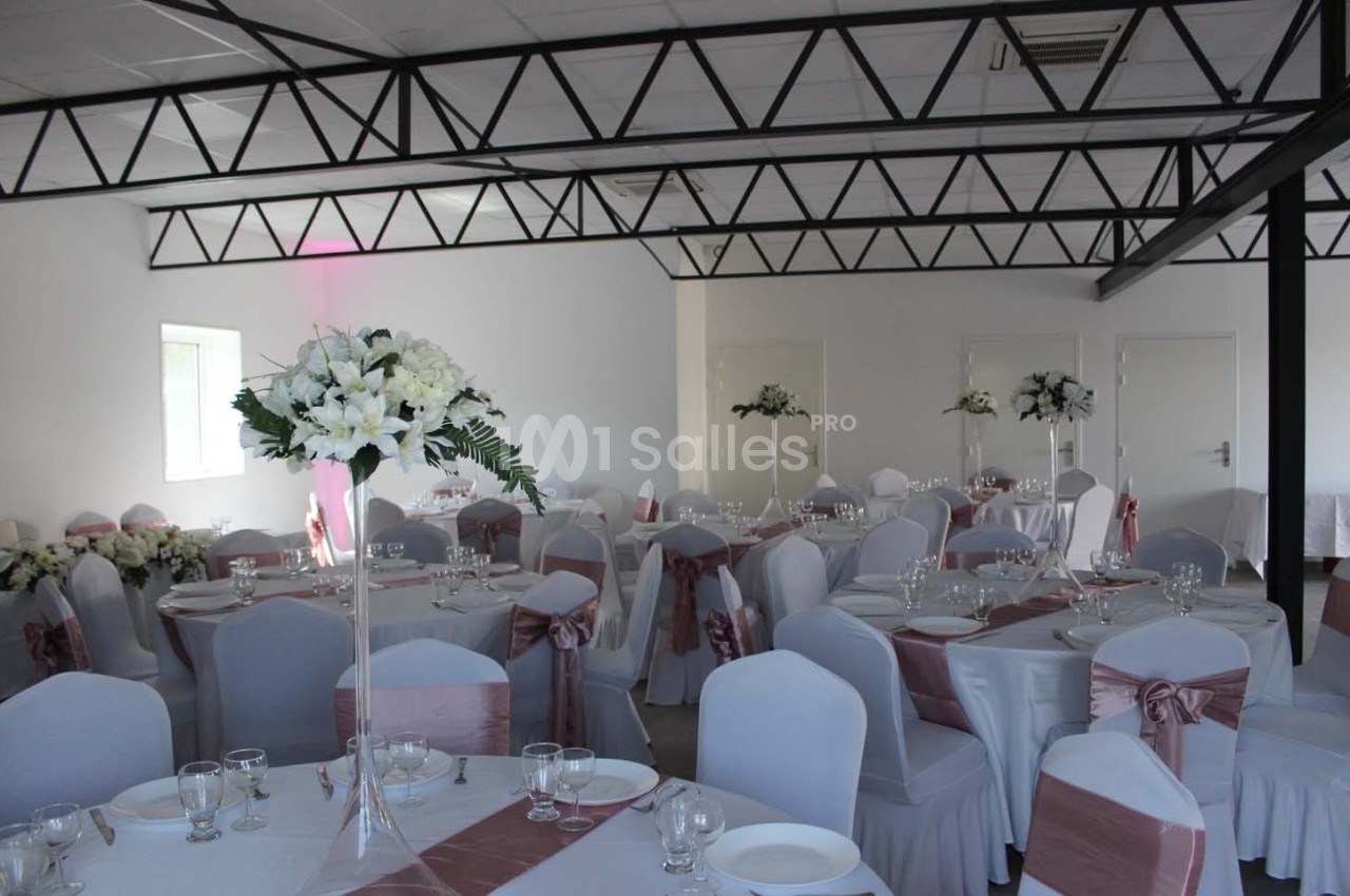 Salle de réception décorée avec des tables rondes, nappes blanches, chaises ornées de nœuds roses et centres de table…