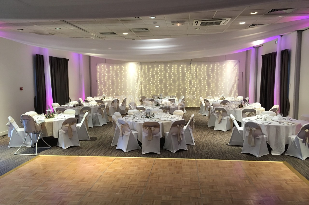 Salle de réception décorée avec des tables rondes dressées et un mur illuminé par des guirlandes lumineuses.