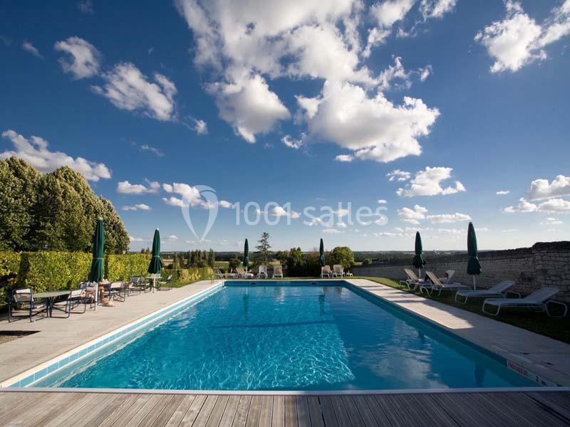 Piscine extérieure entourée de chaises longues, parasols verts et haies, sous un ciel bleu parsemé de nuages.