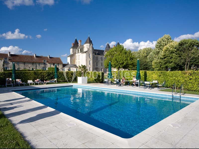Piscine extérieure entourée de chaises longues, avec un château en arrière-plan sous un ciel bleu.