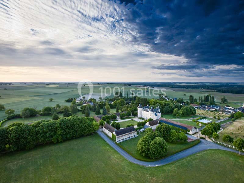 Vue aérienne d'un château entouré de jardins verdoyants et de champs sous un ciel nuageux.