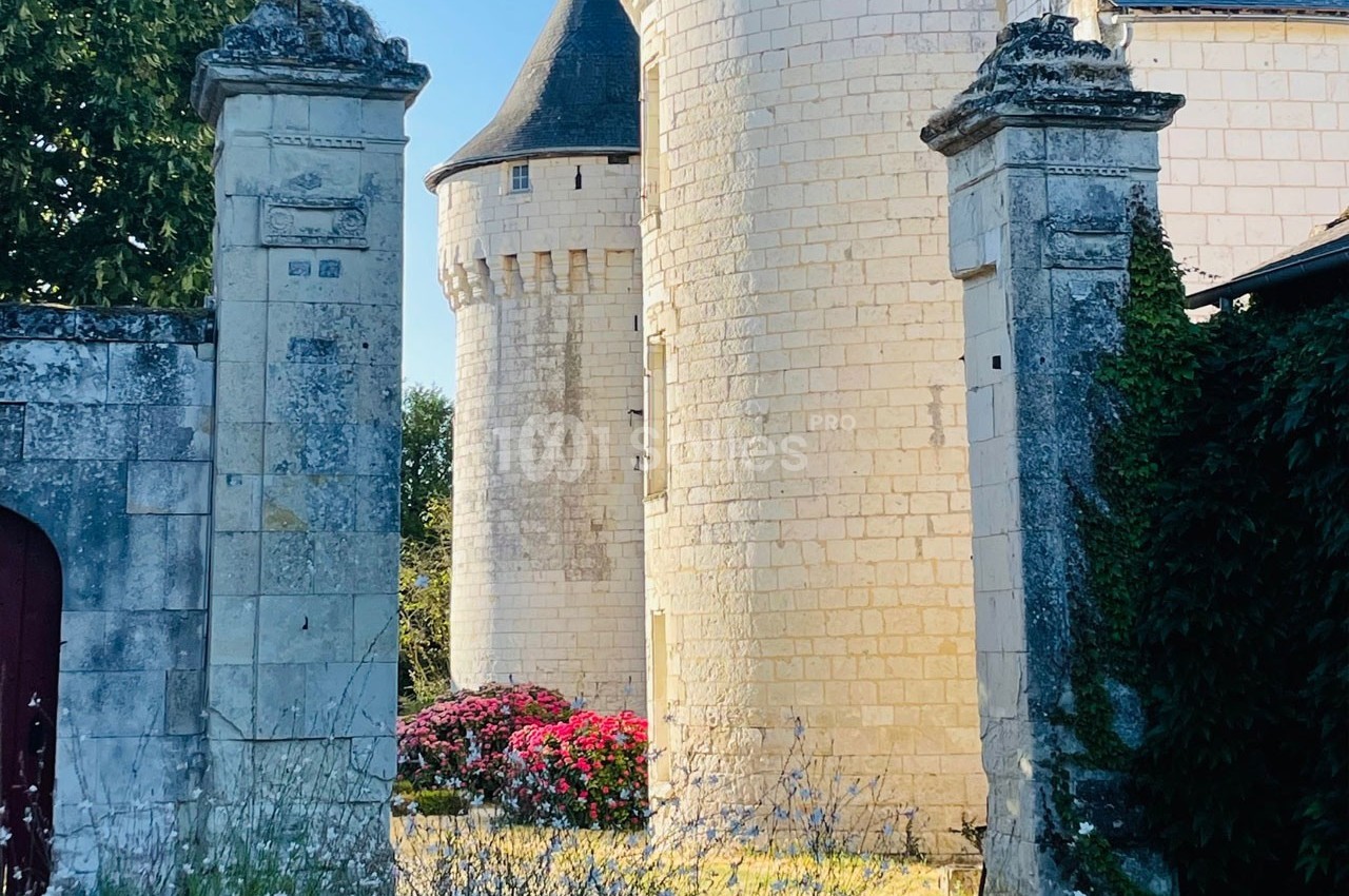 Vue partielle d'un château en pierre blanche avec tours rondes, entouré de végétation et de fleurs colorées.