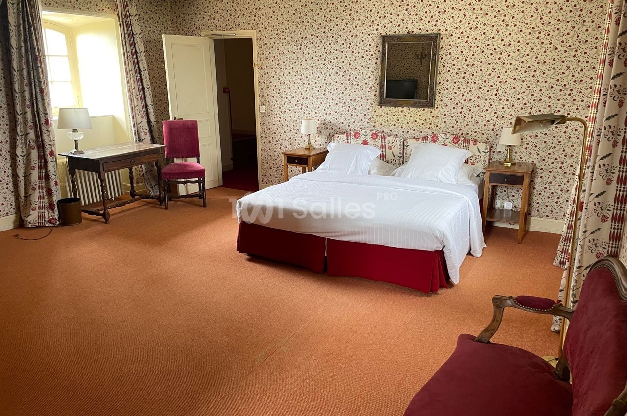 Chambre avec lit double, murs tapissés de motifs floraux, mobilier en bois et fauteuils rouges sur moquette orange.