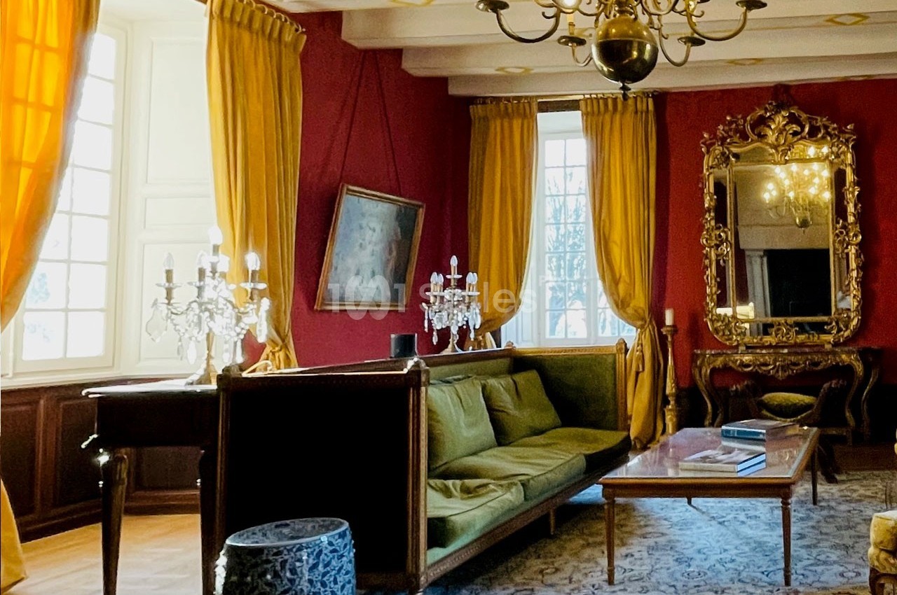 Salon élégant avec murs rouges, rideaux dorés, canapé vert, lustres, miroir doré et lumière naturelle.