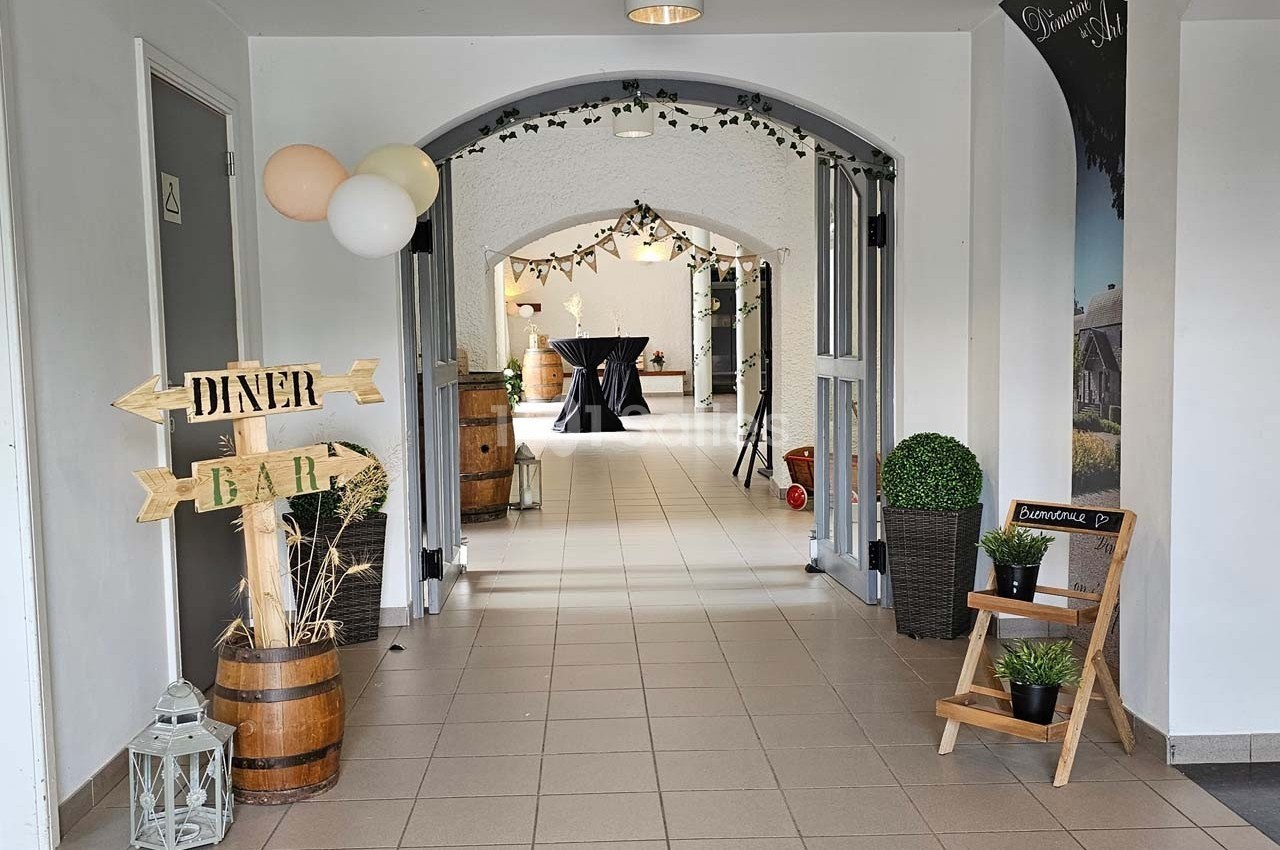 Entrée décorée avec des ballons, panneaux en bois ’Dîner’ et ’Bar’, plantes et tonneaux dans un espace lumineux.