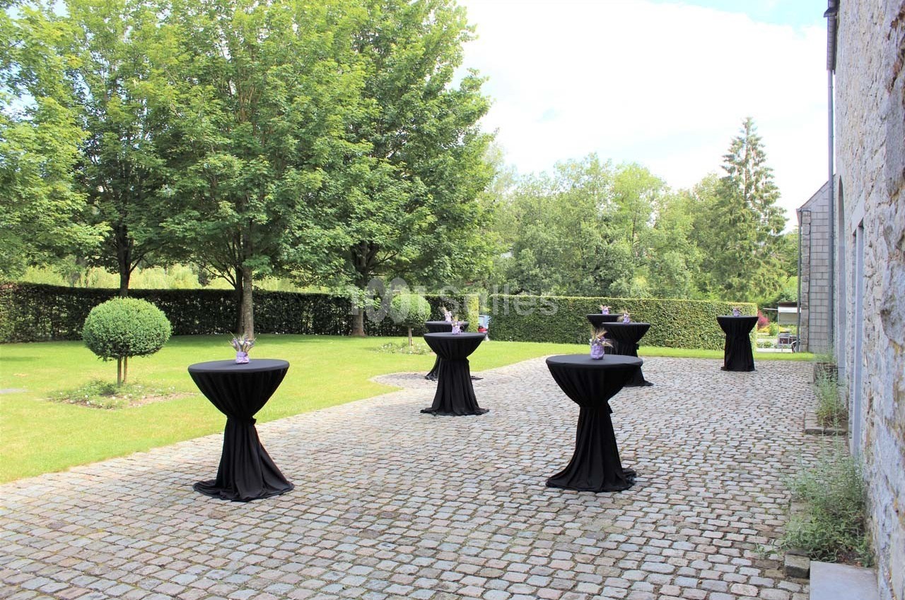 Tables hautes noires disposées sur une terrasse pavée, entourée de verdure et d'arbres.